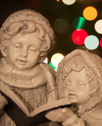 Statue of two small children caroling, Christmas lights out of focus in the background