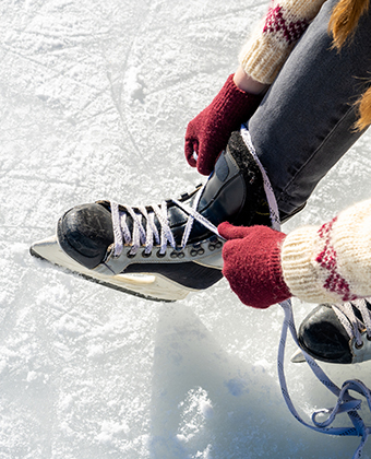 Overhead close up of person wearing a jacket and gloves lacing up ice skates on the ice.